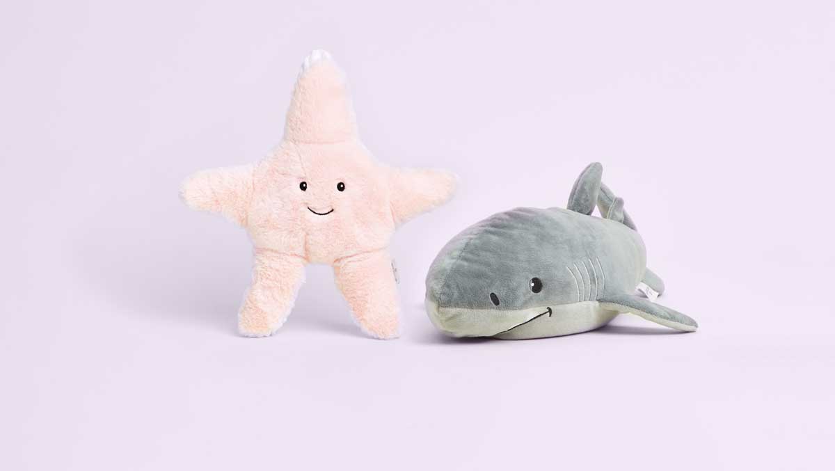 Image with a light pink background featuring a Starfish Warmies and a Shark Warmies.