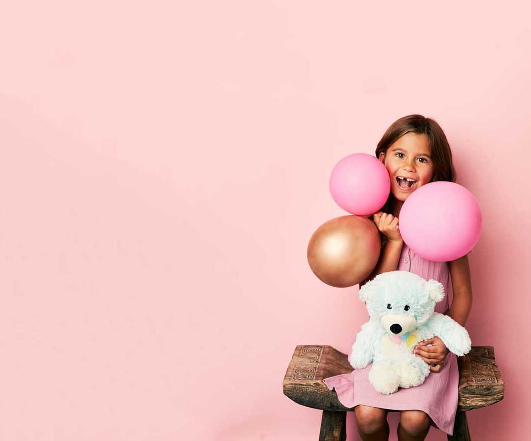 Child smiling holding balloons while holding a Celebration Bear Warmies.